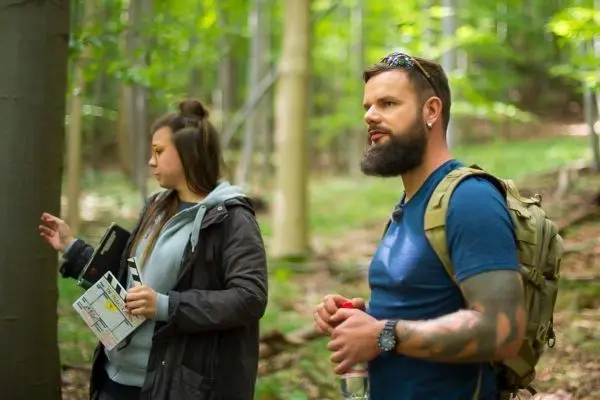Zwei Personen stehen im Wald, eine hält ein Blatt Papier, der andere eine Wasserflasche