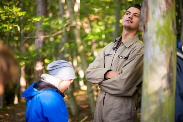 Zwei Personen stehen im Wald, eine lehnt an einem Baum, die andere schaut nach unten