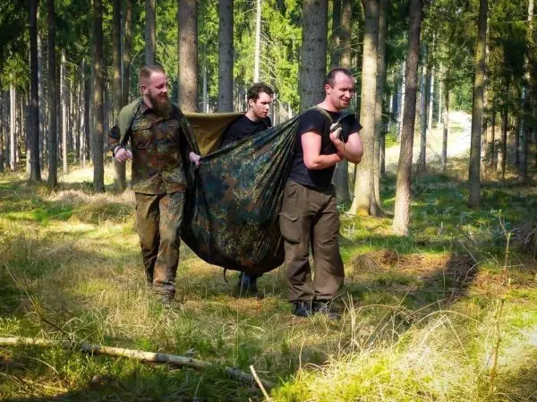 Drei Personen tragen eine improvisierte Trage aus einem Tarp durch den Wald