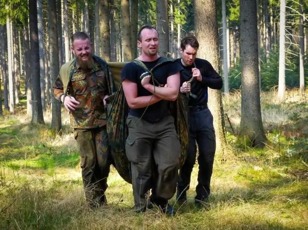 Drei Personen tragen eine improvisierte Trage durch einen Wald