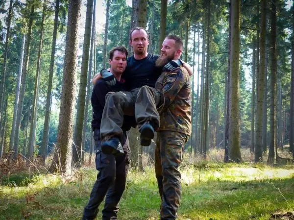 Drei Personen tragen einen Mann durch einen Wald