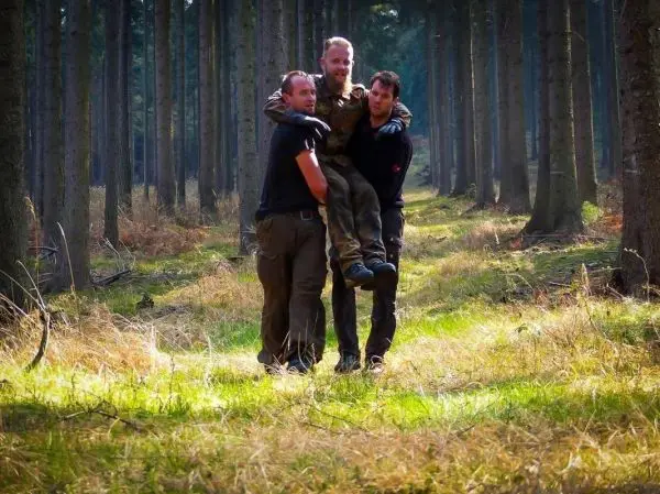 Drei Personen tragen einen Mann durch einen Wald auf einem grasbewachsenen Weg