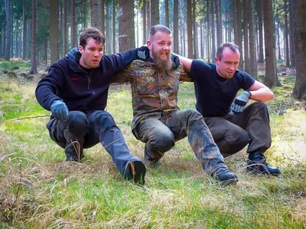 Drei Personen unterstützen sich gegenseitig im Wald, einer wird getragen