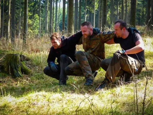 Drei Personen unterstützen sich im Wald beim Tragen eines anderen