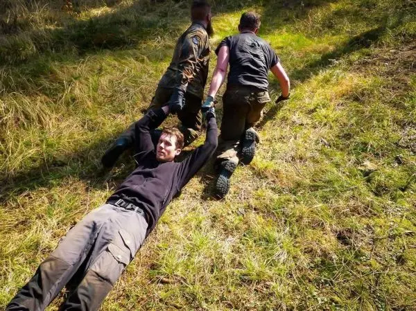 Drei Personen ziehen einen liegenden Mann über grasbewachsene Fläche