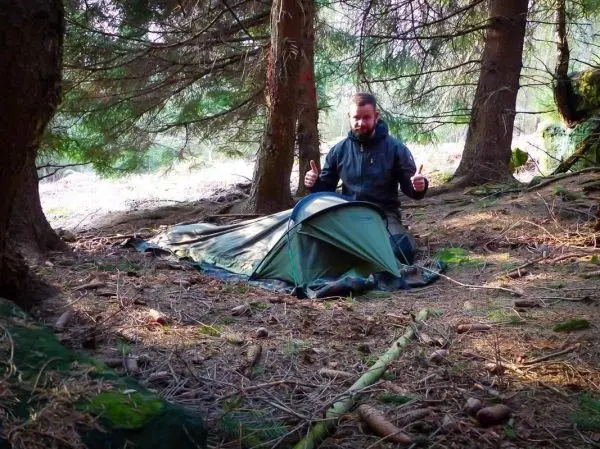 Mann sitzt neben einem aufgebauten Zelt im Wald und zeigt Daumen hoch