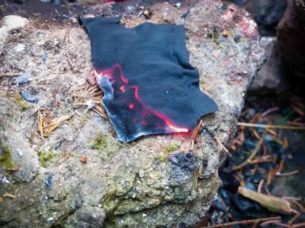 Schwarzes Tuch mit Brandspuren liegt auf einem Stein neben Glutresten