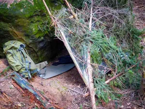 Tarp-Notunterkunft aus Ästen und Zweigen neben einem Felsen, Rucksack liegt daneben