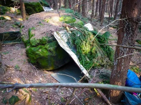 Tarp-Notunterkunft aus Alufolie und Ästen unter einem großen Stein im Wald