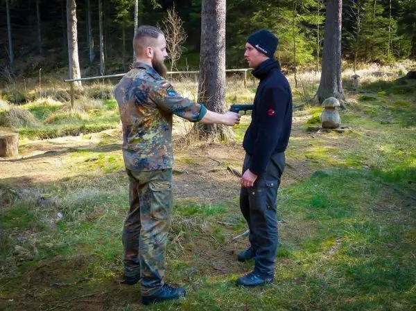 Zwei Personen in einem Wald, eine hält eine Pistole auf die andere