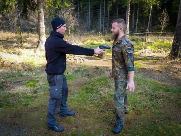 Zwei Personen in einem Wald, eine hält eine Pistole auf die andere gerichtet