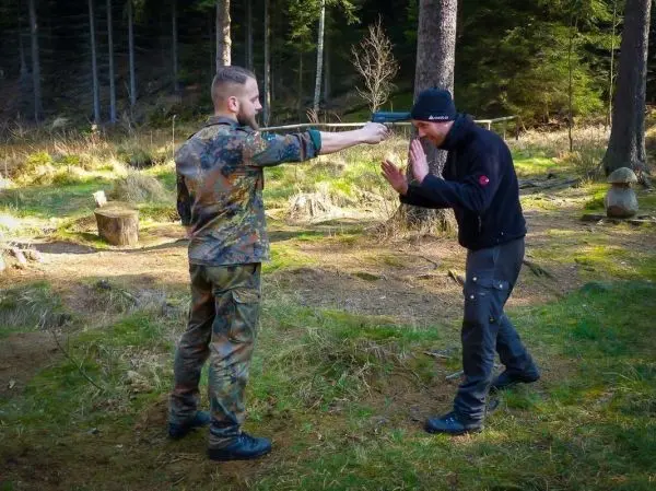 Zwei Personen in einem Wald, eine Person zeigt mit einem Messer auf die andere