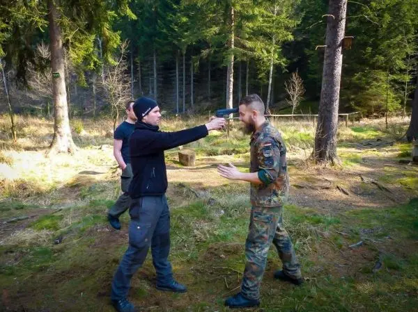 Zwei Personen in militärischer Kleidung und schwarzem Outfit interagieren im Wald