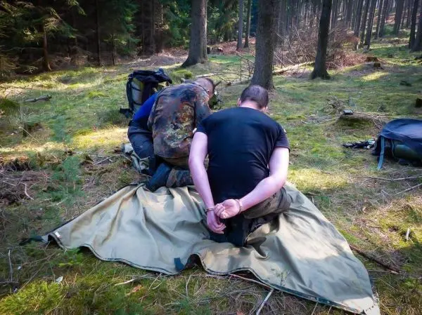 Zwei Personen knien auf einer Tarp-Notunterkunft im Wald, eine ist gefesselt