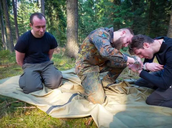 Zwei Personen sitzen auf einer Tarp-Notunterkunft, eine Person fesselt die Hände des anderen