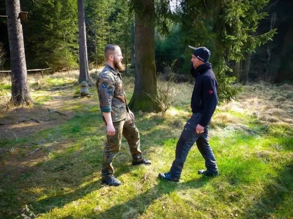 Zwei Personen stehen sich in einem Wald gegenüber, umgeben von Bäumen und Gras