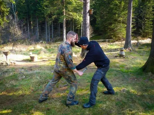 Zwei Personen üben eine Nahkampftechnik im Wald auf einer grünen Wiese