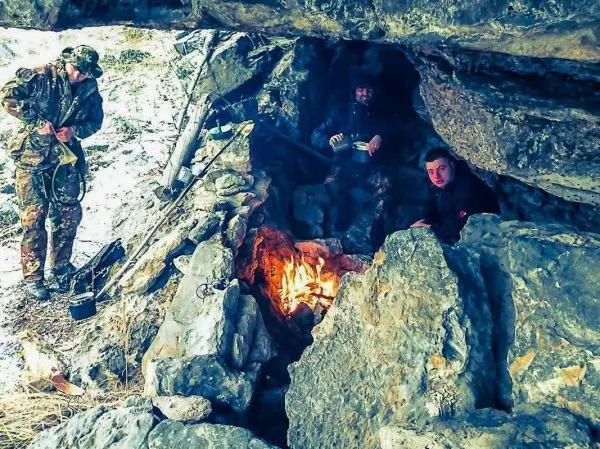 Drei Personen sitzen in einer Felsunterkunft neben einem kleinen Feuer