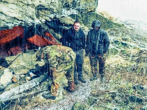 Drei Personen stehen unter einem Felsen, während es regnet. Eine Person beugt sich nach vorne