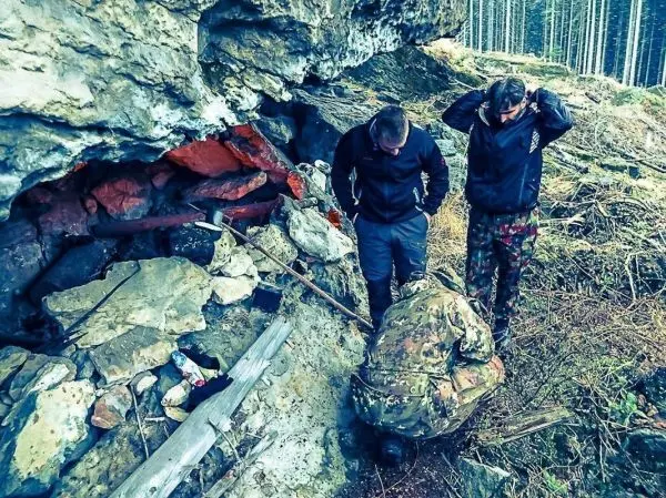 Drei Personen untersuchen eine Felsnische mit Holz und Ausrüstung in einem Waldgebiet
