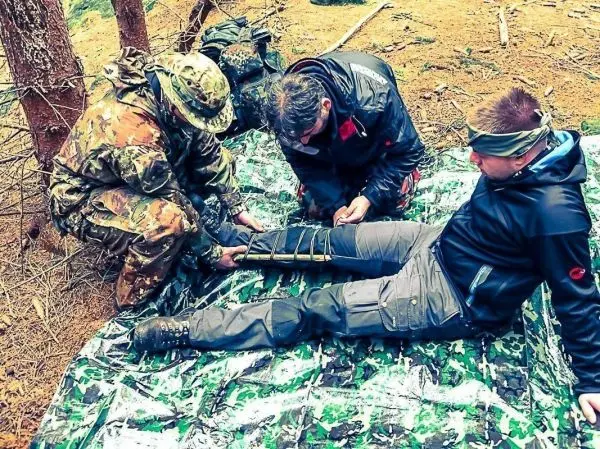 Drei Personen verbinden ein Bein mit improvisiertem Material auf einer camouflierte Unterlage