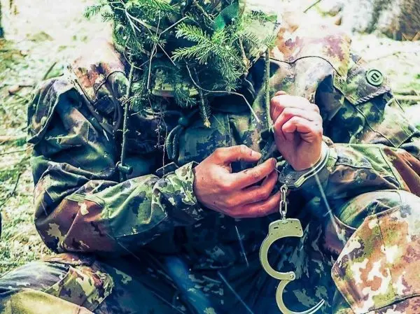 Hände mit Handschellen in Tarnkleidung, umgeben von Zweigen und Laub