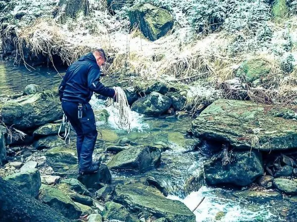 Mann hält ein Seil über einem fließenden Gewässer zwischen großen Steinen