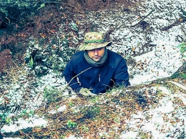 Mann in dunkler Jacke und Hut sitzt am Rand eines bewaldeten Hangs