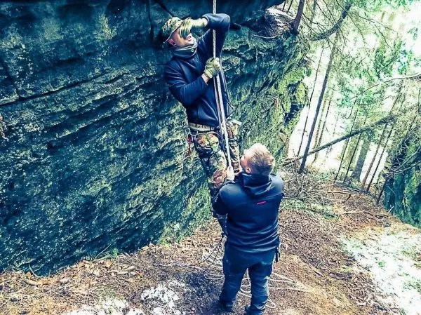 Mann zieht sich an einem Seil an einer Felswand hoch, während ein anderer Mann hilft