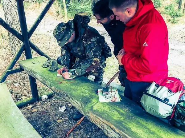 Person zeichnet auf Holzplatte, zwei Personen schauen zu beim Überlebenstraining