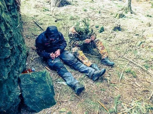 Zwei Personen sitzen am Boden, eine bearbeitet Holz mit einem Messer