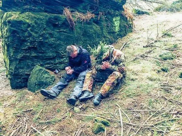 Zwei Personen sitzen an einer Felswand im Wald und halten ihre Hände zusammen