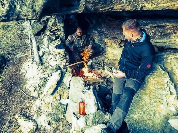 Zwei Personen sitzen in einer Höhle neben einem Feuer, während ein Topf über den Flammen hängt
