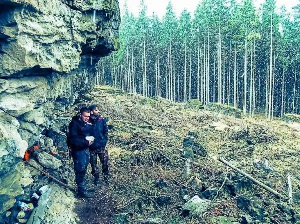 Zwei Personen stehen an einer Felswand im Wald und halten einen Behälter