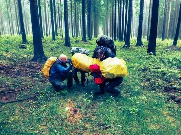 Drei Personen mit Rucksäcken helfen beim Anheben eines anderen Rucksacks im Wald