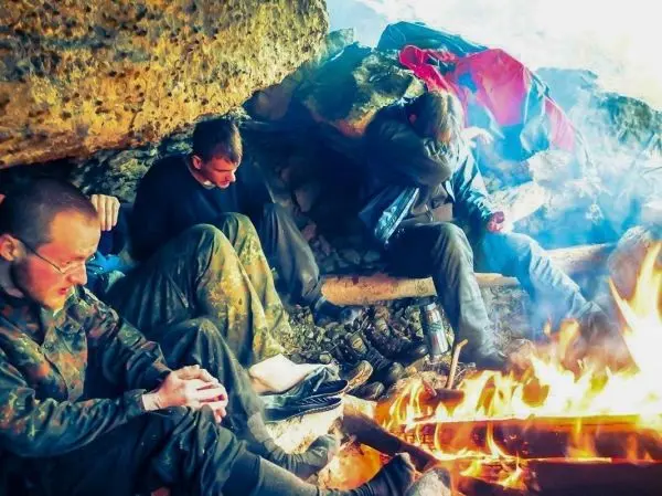 Kleine Flamme brennt vor einer Felsunterkunft, drei Personen sitzen im Hintergrund