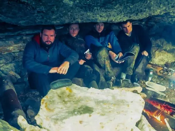 Vier Personen sitzen unter einem Felsvorsprung, ein kleines Feuer brennt vor ihnen