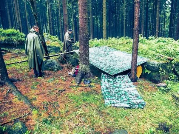 Zwei Personen bauen eine Tarp-Notunterkunft zwischen Bäumen im Wald auf