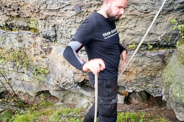 Mann hält einen Stock und steht vor einer Felswand mit Pflanzenbewuchs
