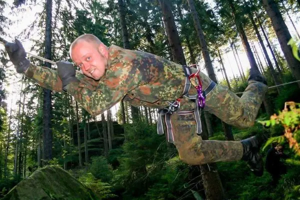 Mann in Tarnuniform überquert selbstgebaute Seilbrücke zwischen Bäumen