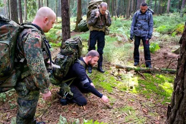 Mann mit Rucksack untersucht den Boden im Wald, während andere zuschauen