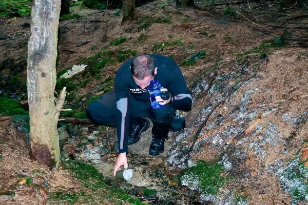 Person beugt sich über einen kleinen Bach und hält eine Wasserflasche in der Hand