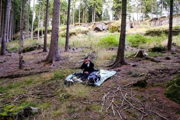 Person sitzt auf einer Tarp-Notunterkunft im Wald und hält einen Gegenstand in der Hand