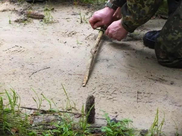 Zwei Hände formen eine Kerbe in einen Holzstock auf dem Boden