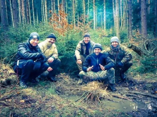 Fünf Personen posieren lächelnd im Wald neben einem kleinen Feuer