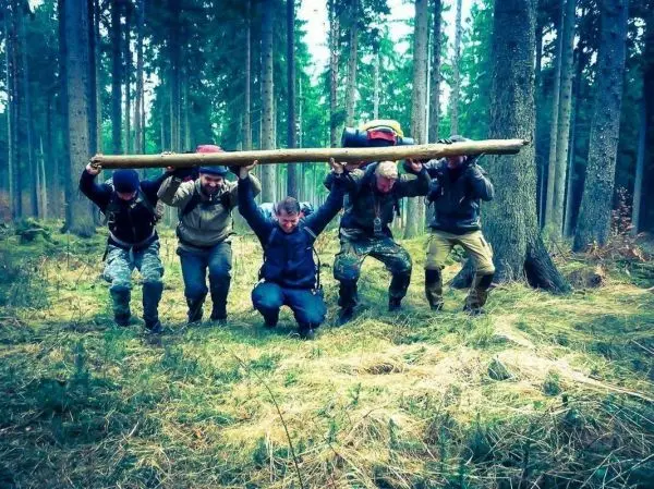 Fünf Personen tragen gemeinsam einen langen Holzstock über den Schultern im Wald