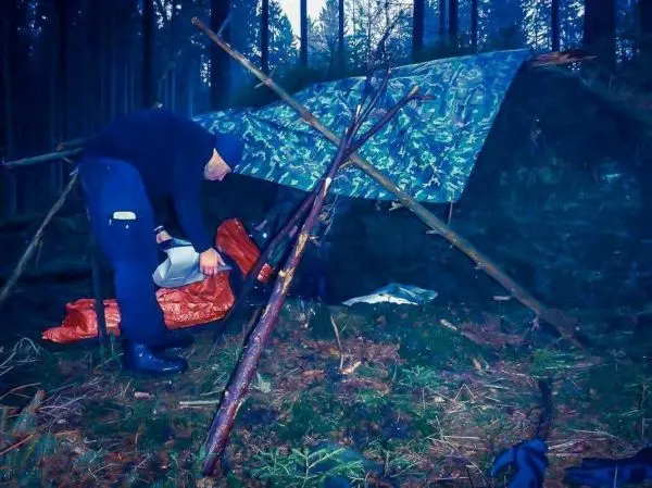 Mann richtet Tarp-Notunterkunft mit Holzstöcken und orangefarbener Plane ein