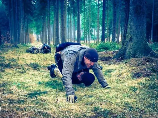 Person krabbelt auf dem Boden im Wald, weitere Personen im Hintergrund