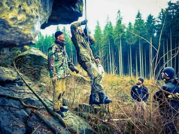 Person zieht sich an einem Seil hoch, andere beobachten aufmerksam beim Überlebenstraining