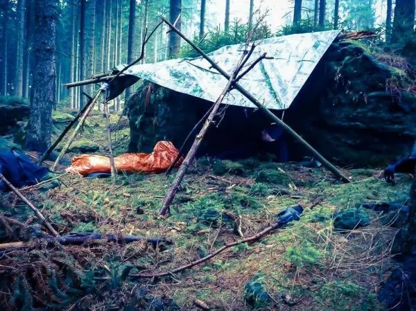 Tarp-Notunterkunft aus Ästen und Plane neben einem Felsen im Wald